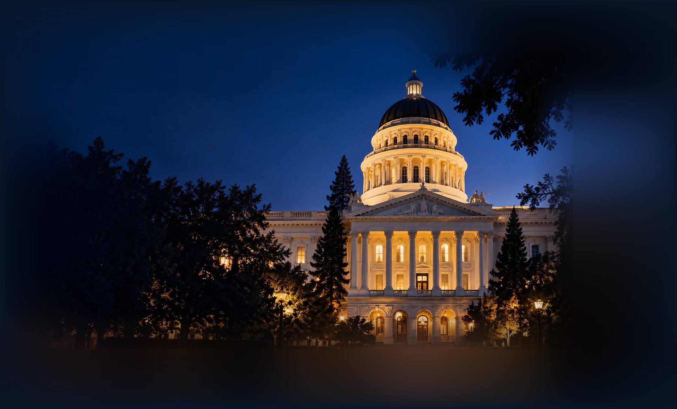 The California State Capitol at night, its dome and columned entrance warmly lit against a deep blue sky and framed by dark trees.